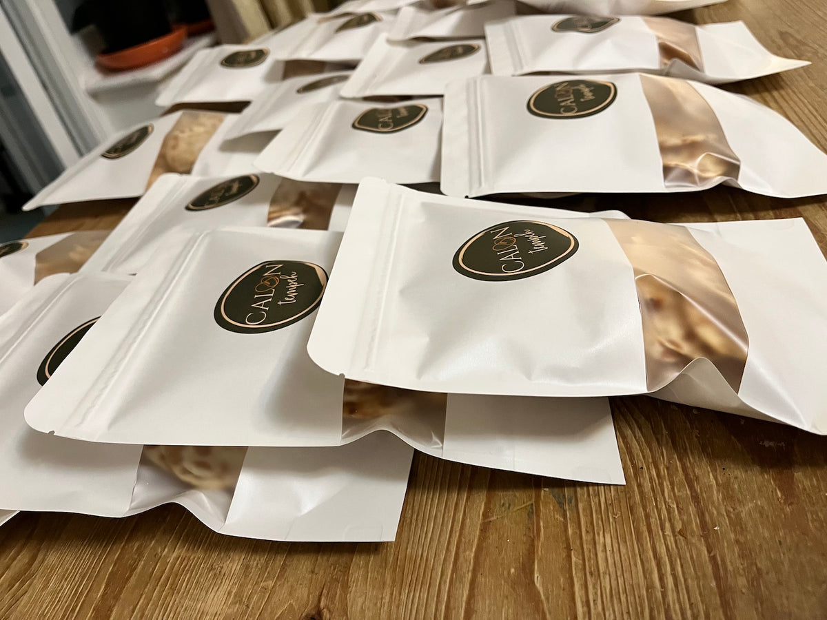 Multiple white resealable stand-up pouches of Calon Tempeh Crisps with oval dark green-and-gold labels are arranged on a wooden tabletop. The front pouch is most in focus, with several identical pouches blurred in the background. Contains tempeh crisps / tempe chips.