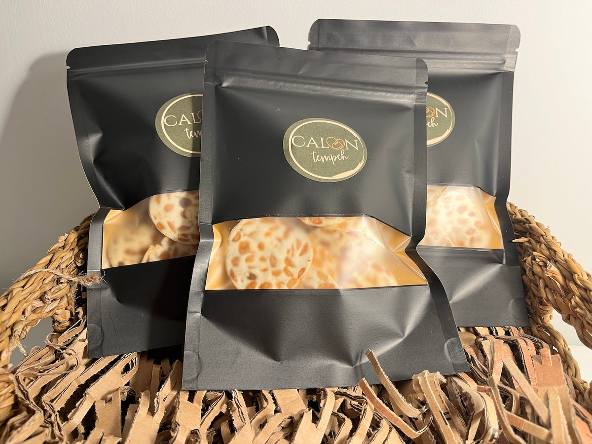 Three black stand-up pouches of Calon Tempeh Crisps/ tempe chips arranged in a woven basket. Each pouch has a circular gold logo and a clear window showing the round tempeh crisps/  tempe chips inside.