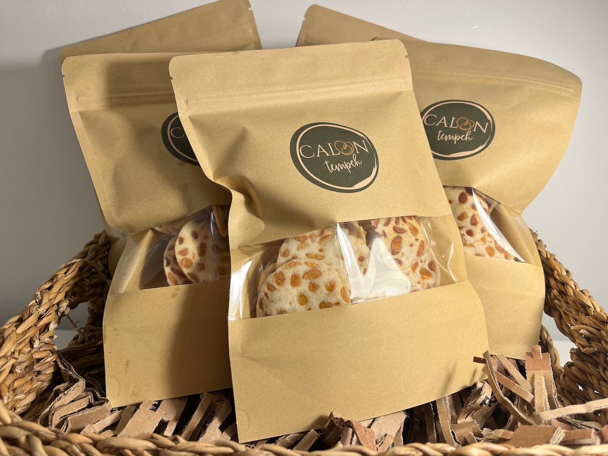 Three kraft paper stand-up pouches of Calon Tempeh crisps / tempe chips , with the center pouch open to reveal tempeh crisps/ tempe chips  inside, arranged in a woven wicker basket on shredded paper.