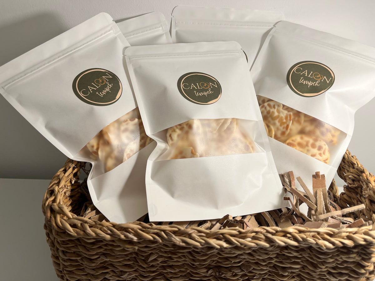 Calon Tempeh Crisps / tempe chips Welsh Salt. Three white stand-up tempeh crisps pouches with clear windows displaying  tempeh crisps / tempe chips , arranged in a woven wicker basket with shredded paper. Each pouch features a circular gold and dark green logo reading 'Calon Tempeh'.