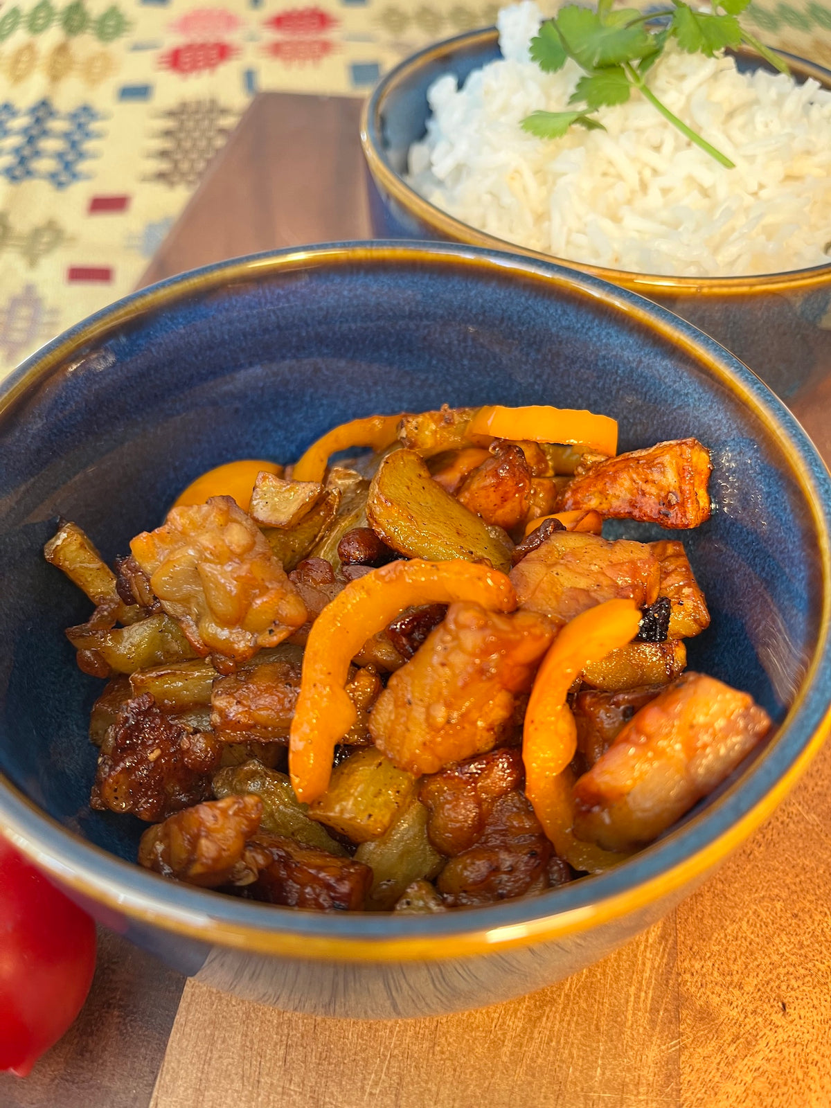 A close-up shot of a blue ceramic bowl filled with a stir-fried crispy tempeh mixed with Calon Tempeh sweet chilli sauce and vegetable  served with a side of white rice in another bowl on a wooden table.