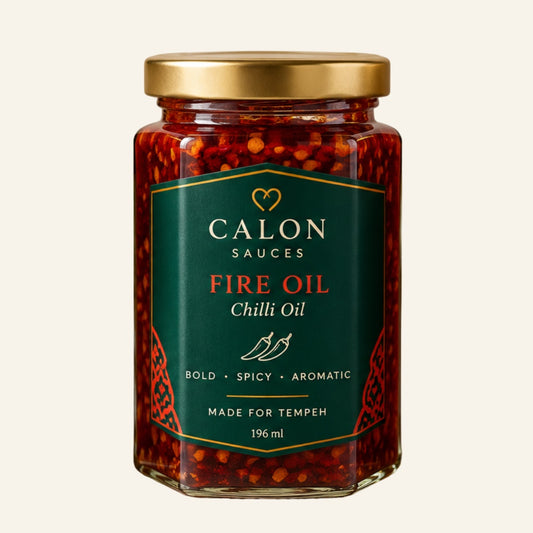 A glass jar of Calon Tempeh Sauces Fire Oil Chilli Oil with a gold lid and a dark green label, set against a solid off-white background. Perfect for cooking with fresh tempeh.