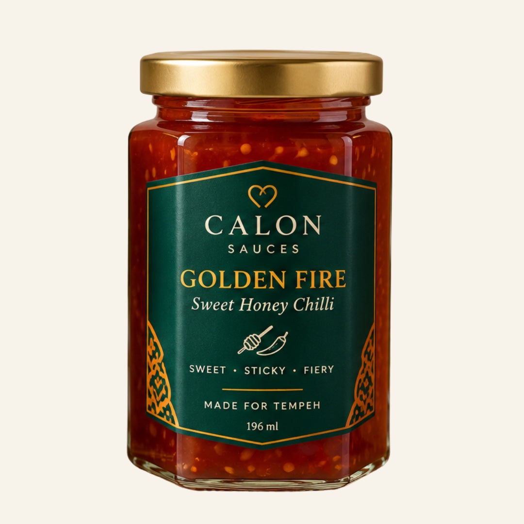 A glass jar of Calon Tempeh Sauces Golden Fire Oil, sweet honey chilli sauce,  with a gold lid and a dark green label, set against a solid off-white background. Perfect for cooking with fresh tempeh.