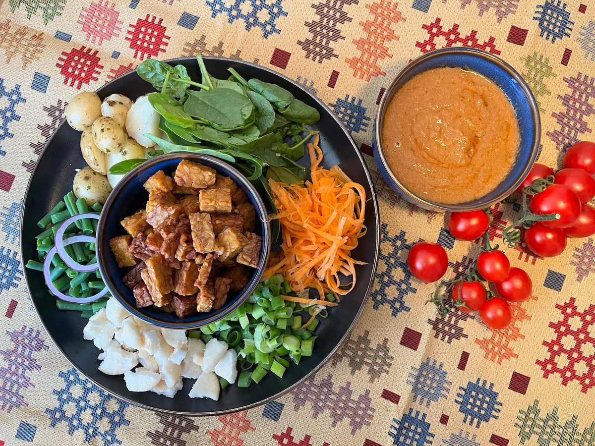 A styled overhead shot of a prepared food dish,  a vegan tempeh salad, served on a black plate with various colourful ingredients including spinach, potatoes, onions, carrots, and green onions, accompanied by a side of Calon Satay sauce and fresh cherry tomatoes on a Welsh-inspired patterned tablecloth.