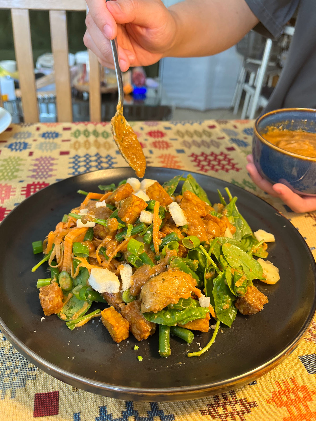 A close-up of a prepared food dish on a black plate, featuring cooked greens, tofu, fried tempeh, and pieces of rice crackers, with a hand holding a spoon adding satay sauce. A bowl is held on the right, set on a Welsh-inspired patterned tablecloth in an indoor dining environment.