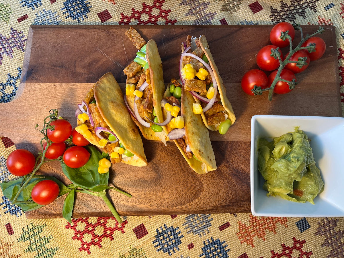 Three stuffed soft corn tacos filled with fried tempeh, corn, red onion, and edamame beans, served on a wooden board with tomatoes and a side of guacamole.